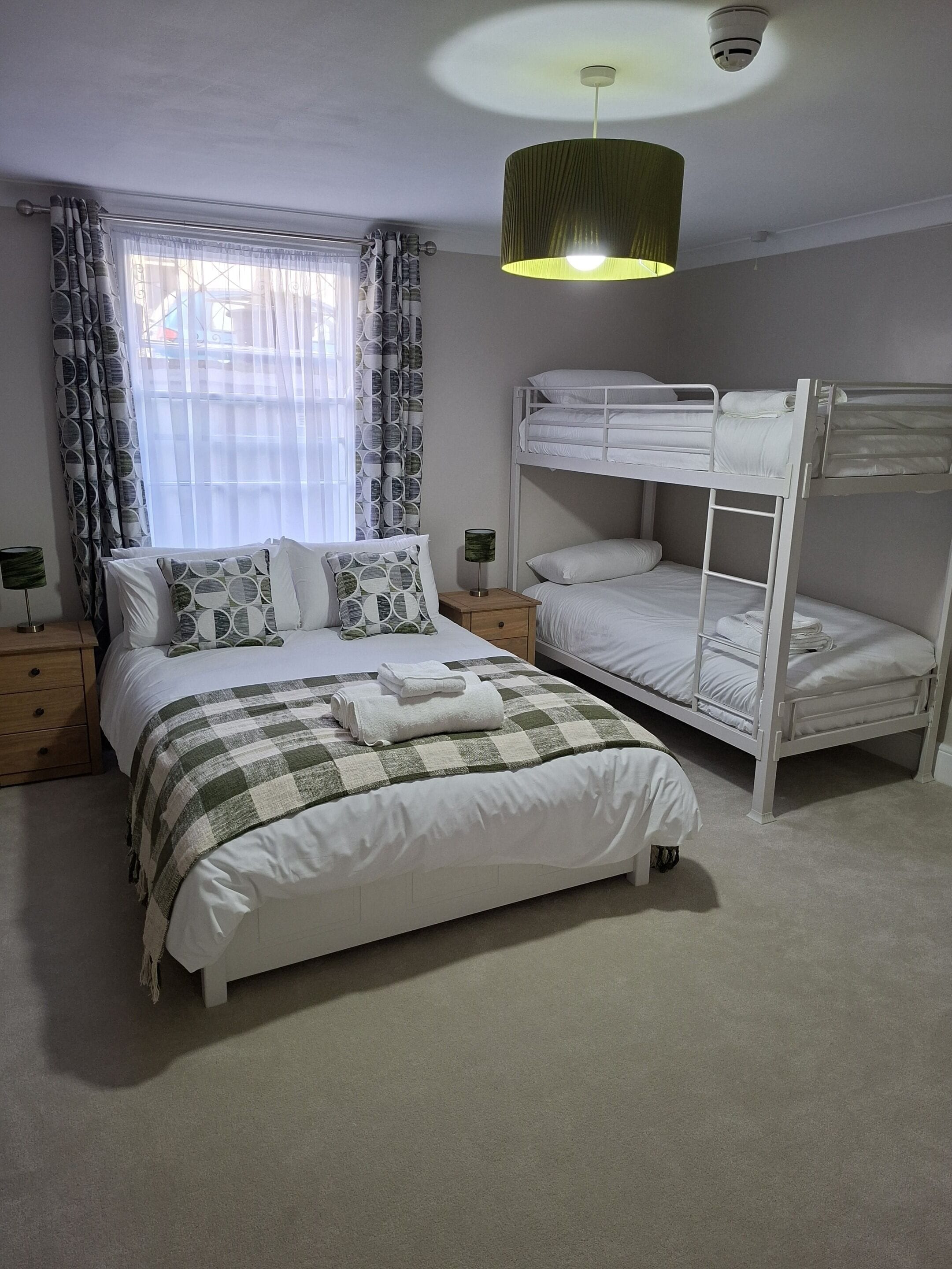Un dormitorio con una cama litera, una cama individual, una mesita de noche con lámpara y una ventana con cortinas.