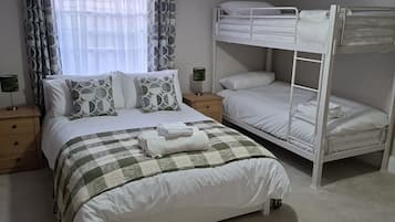 Un dormitorio con una cama litera, una cama individual, una mesita de noche con lámpara y una ventana con cortinas.