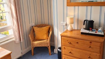 A room with a striped wall, a wicker chair, a wooden dresser, a lamp, and a window with curtains.