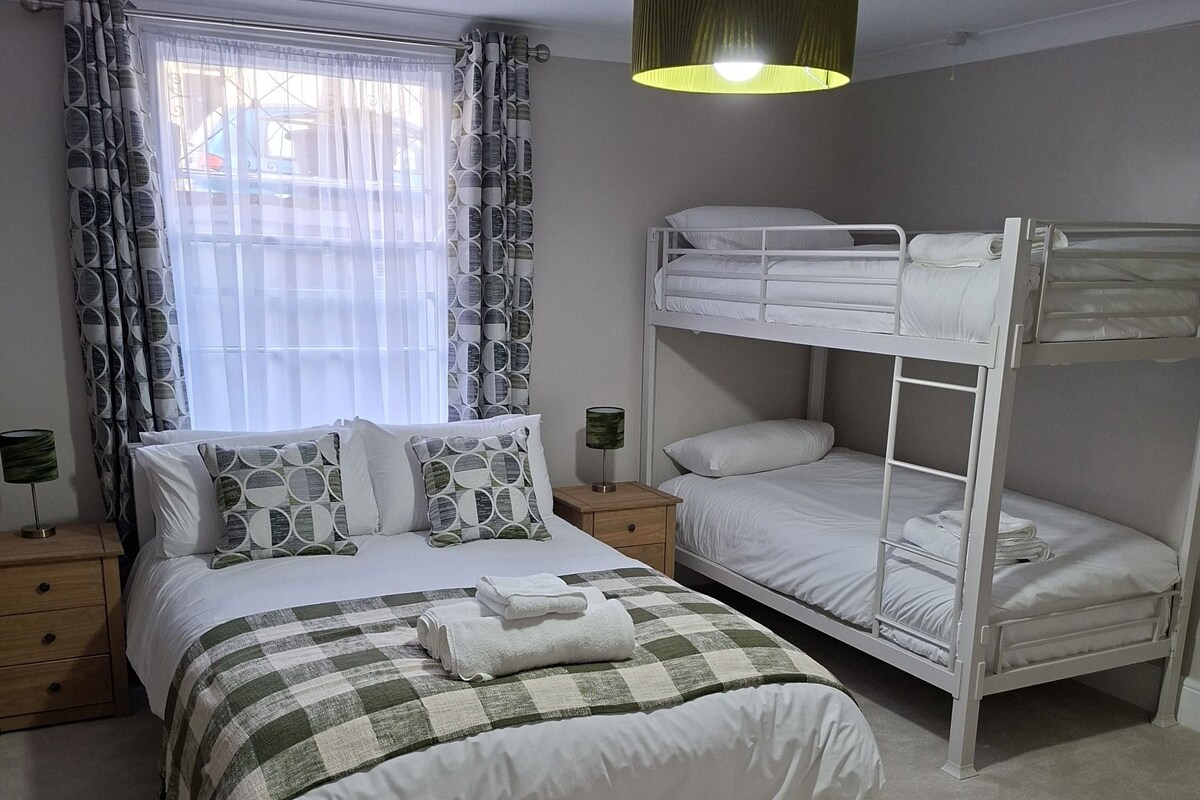 A bedroom with a bunk bed, a single bed, a bedside table with a lamp, and a window with curtains.
