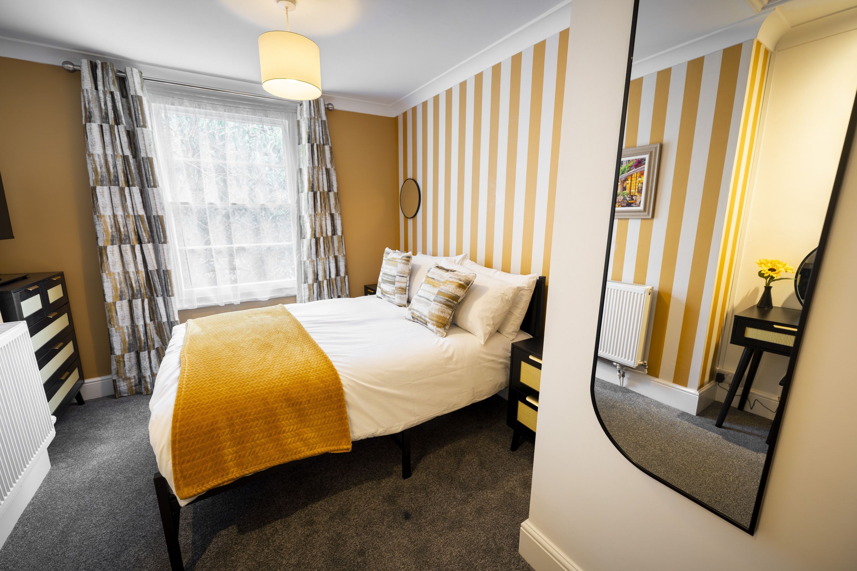 A hotel room with a bed, a bedside table, a mirror, a radiator, and a window with curtains.