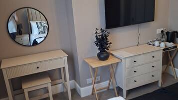 A bedroom with a bed, a desk, a chair, a TV, a small table with a plant, and a bedside table with a lamp.