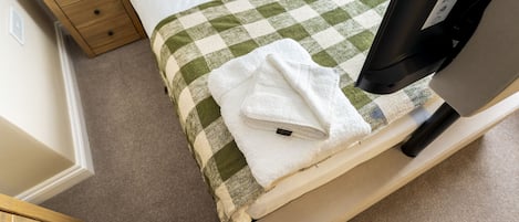 Una cama bien hecha con una colcha a cuadros, una toalla blanca sobre la cama, una mesita de noche de madera con una lámpara, y un televisor negro al lado.