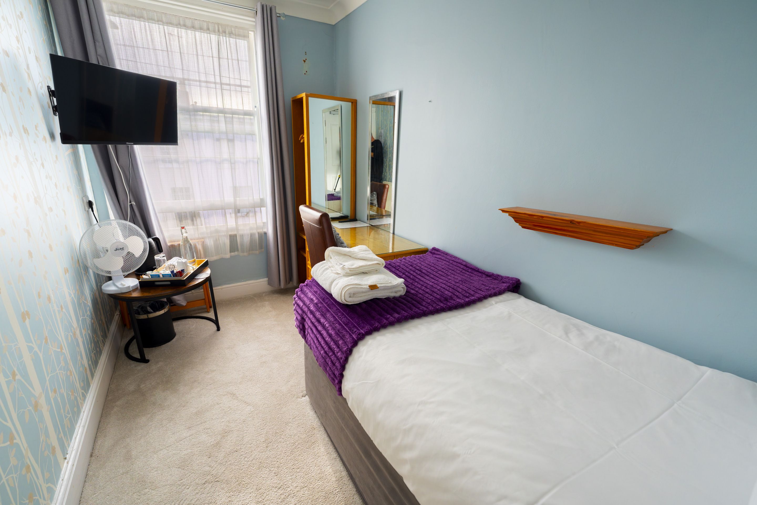 A hotel room with a bed, a desk with a fan, a mirror, and a TV mounted on the wall.