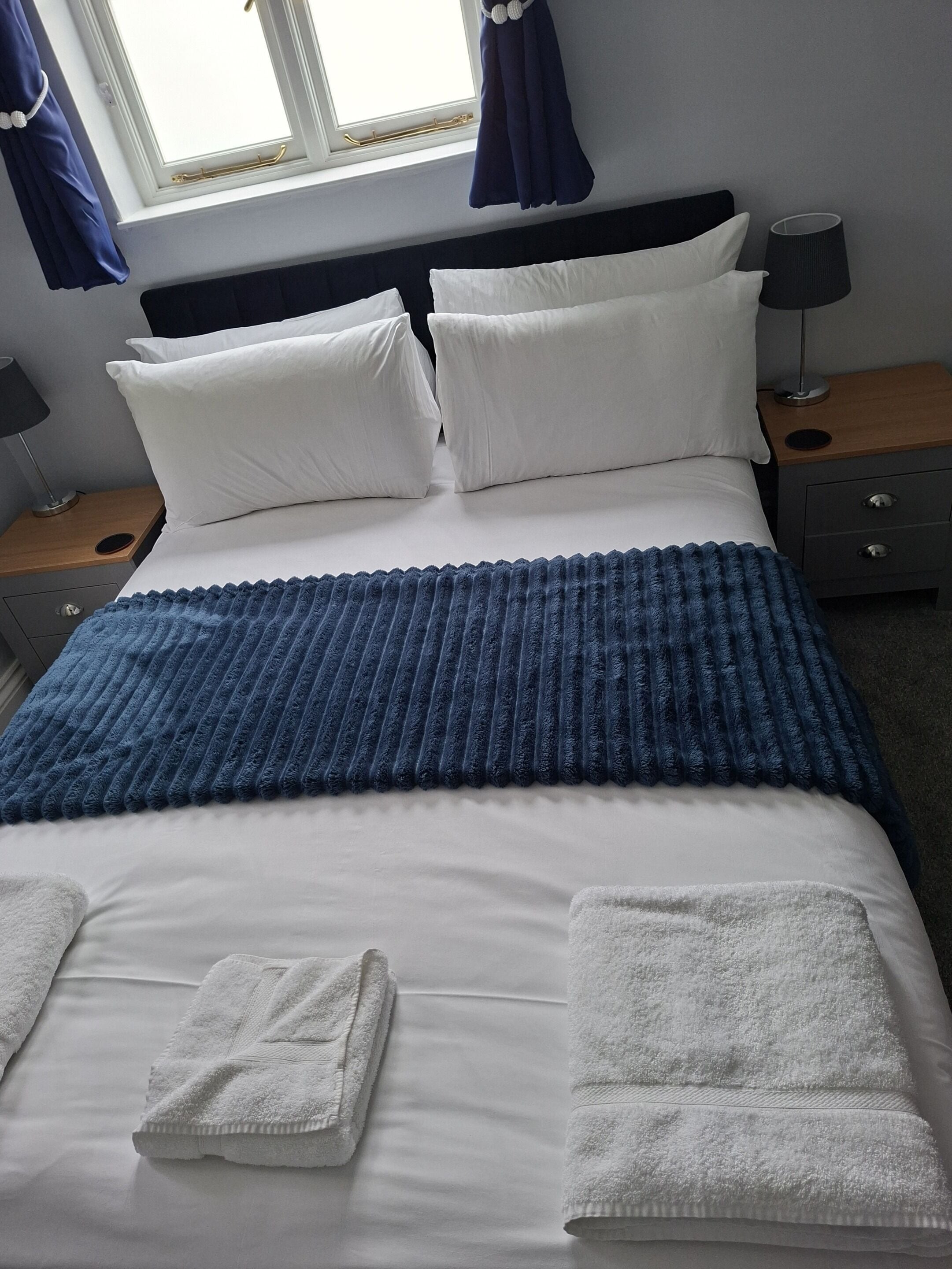 A neatly made bed with white linens and a blue blanket, two bedside tables with lamps, a window with curtains, and a wall-mounted lamp.