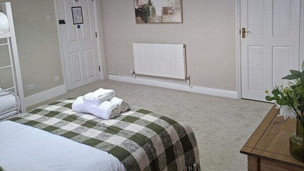 A bedroom with a bed, a bedside table with a vase, a radiator, and a door. - The Abbey (Cheltenham)