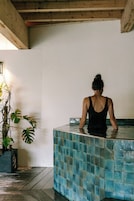 A person in a black tank top standing in a pool with blue tiles, surrounded by indoor plants and wooden beams.