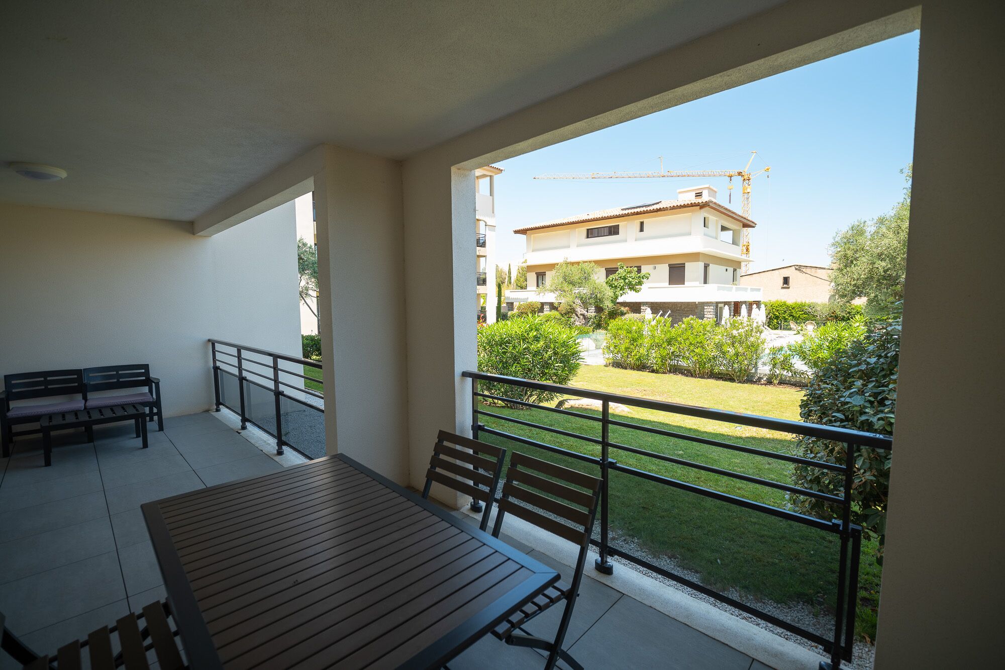 Un balcon avec une table et des bancs, donnant sur une cour verdoyante et un bâtiment en construction.
