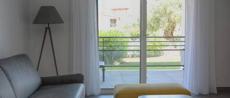 Ein modernes Wohnzimmer mit einem grauen Sofa, einem kleinen gelben Hocker, einer Stehlampe und einer Glasschiebetür, die auf einen Balkon mit Blick auf Grünflächen und ein Gebäude im Hintergrund führt.