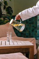 A hand pouring water from a glass jug into a glass on a wooden table.