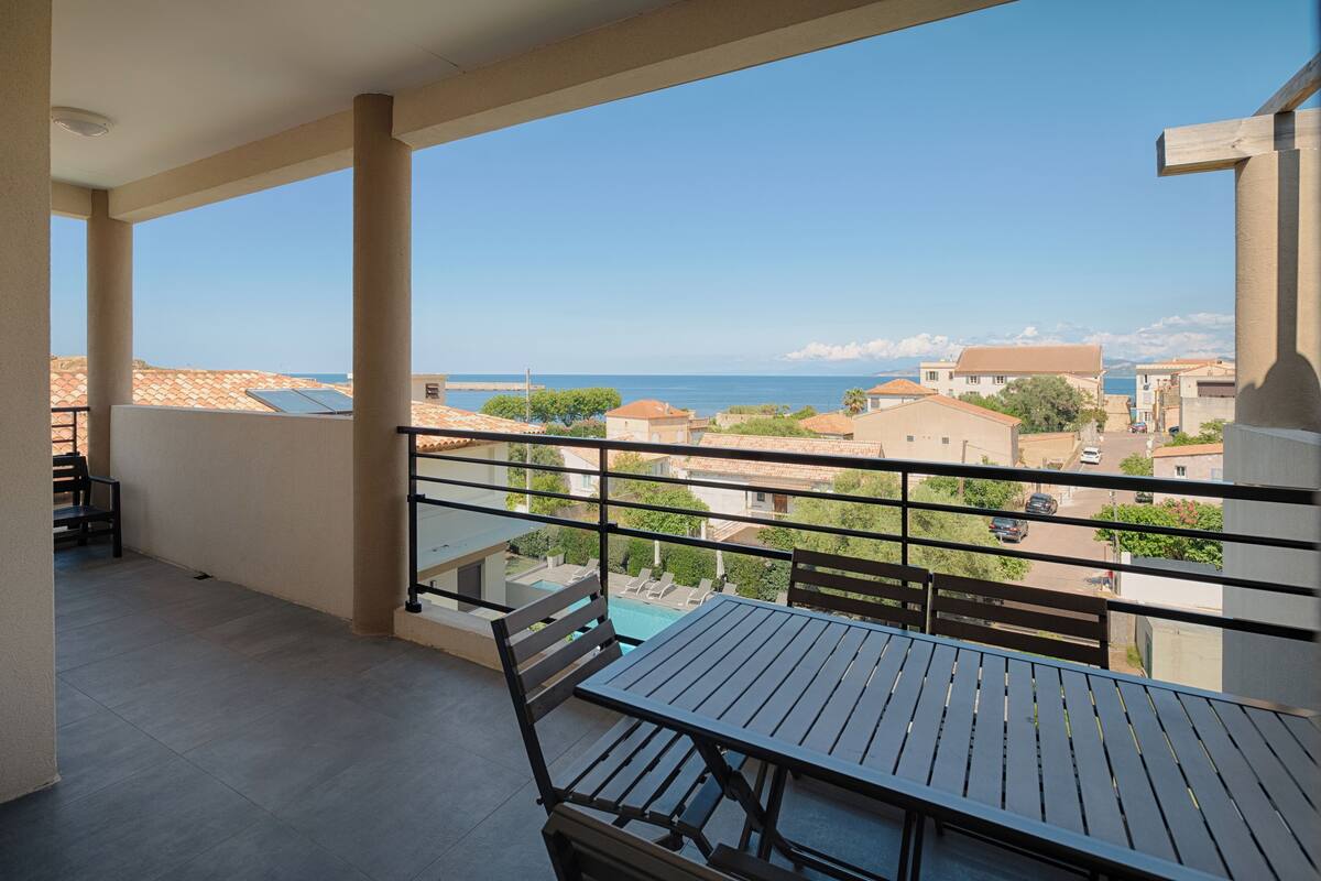 A balcony with a table and chairs, overlooking a residential area and the sea.