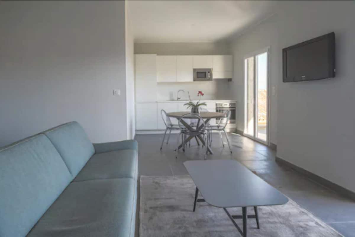 A modern living room with a sofa, a coffee table, and a dining area with a table and chairs. A kitchen with white cabinets and a microwave is visible in the background.