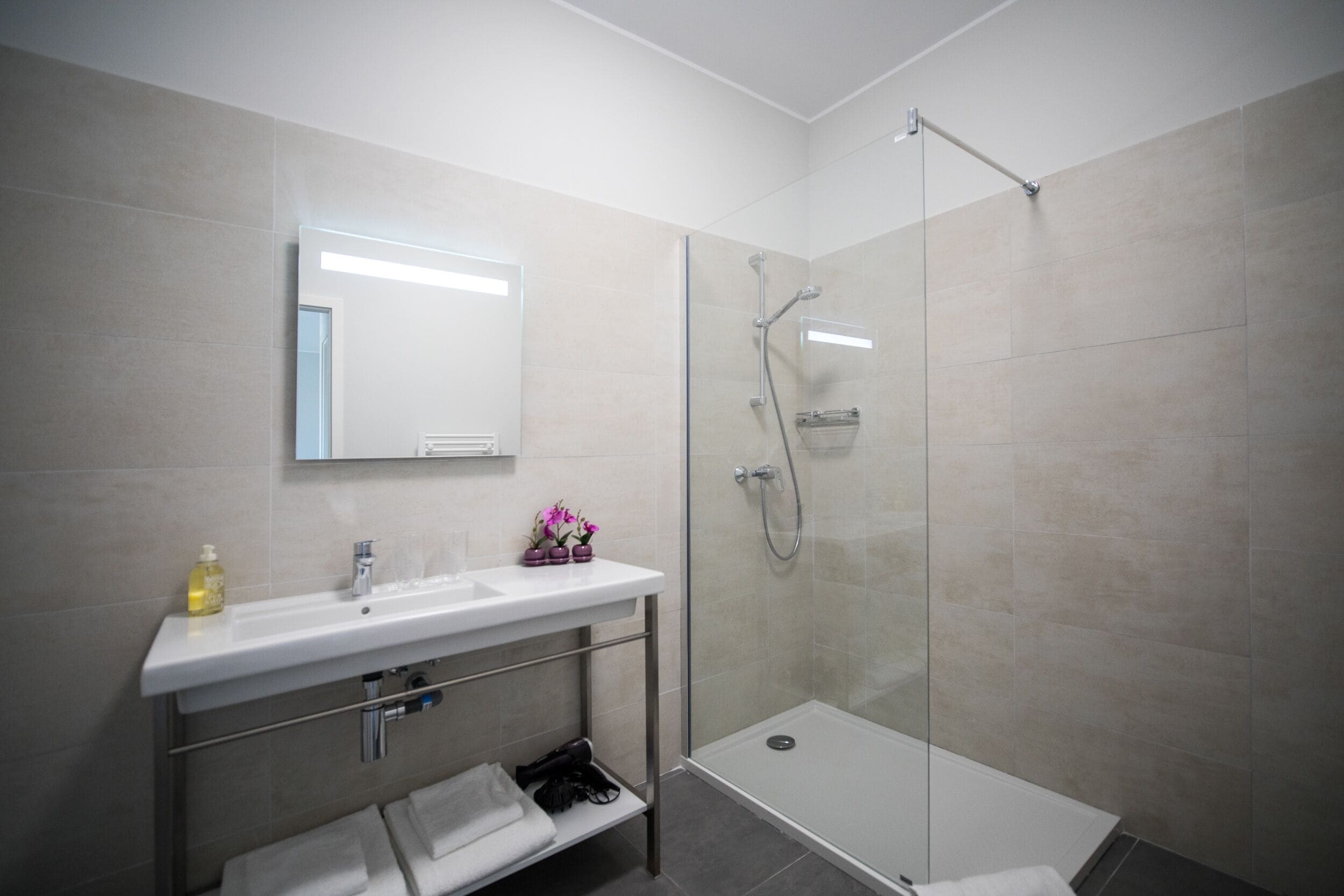 A modern bathroom with a glass shower enclosure, a white sink with a mirror, and a shelf with towels.