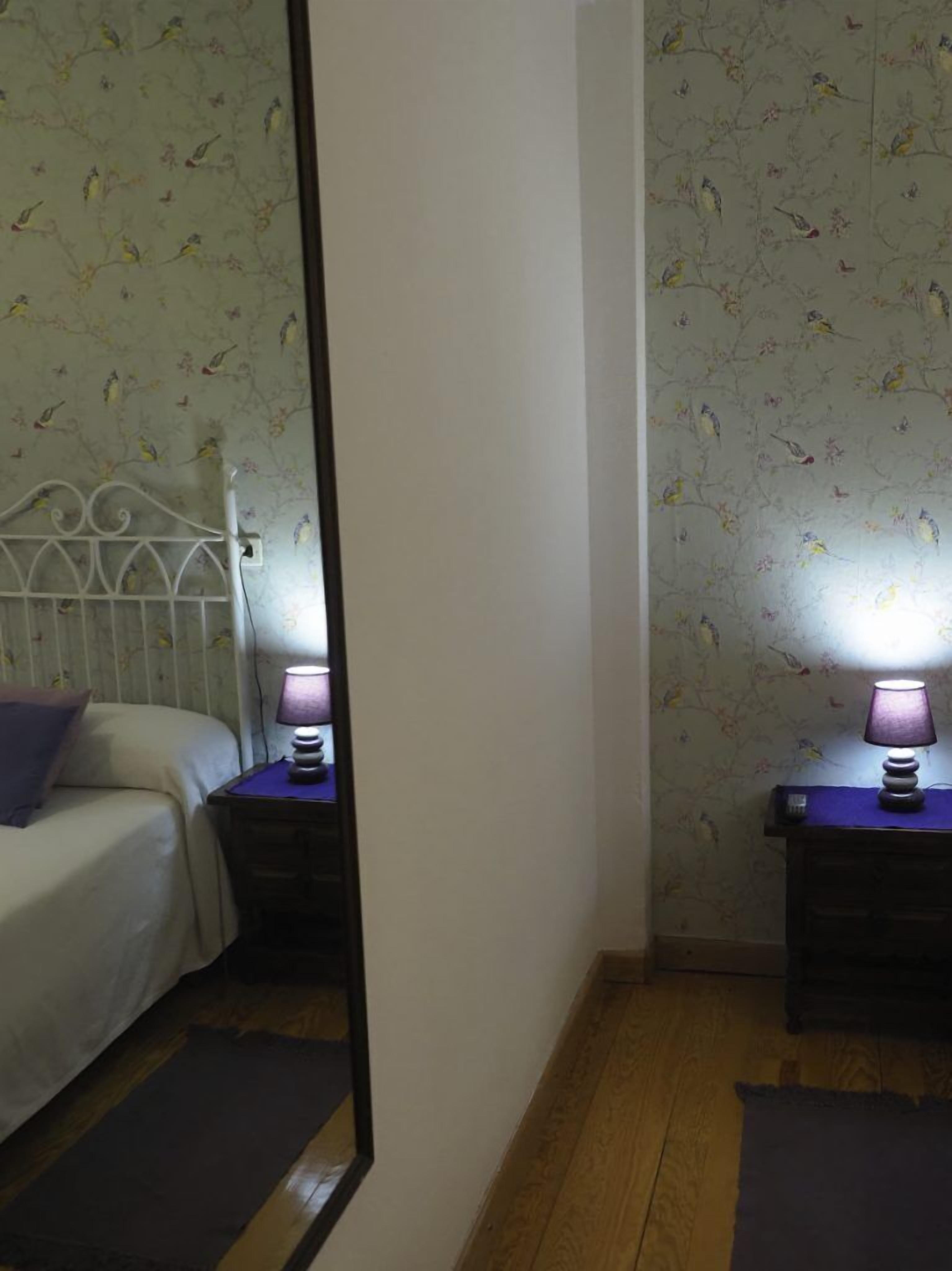 A bedroom with a bed, two bedside tables with lamps, a mirror, and floral wallpaper.