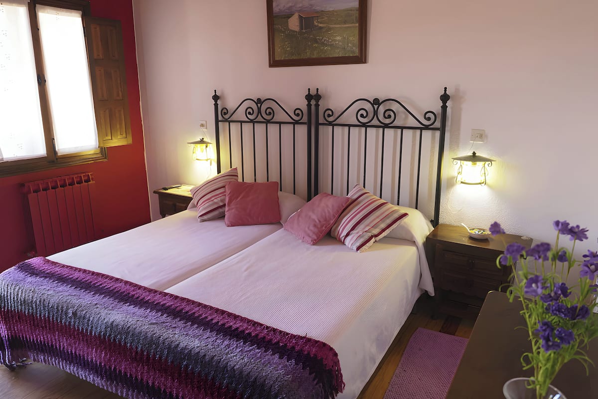 Un lit double avec une couvre-lit à rayures, deux tables de chevet avec des lampes, un tableau représentant un paysage encadré et un radiateur.