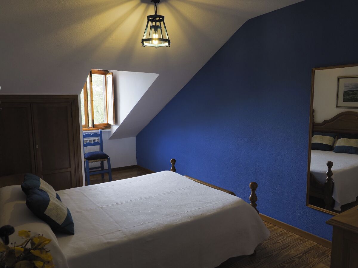 A bedroom with a sloped ceiling, a bed with a wooden headboard, a chair, a desk, and a window with natural light.