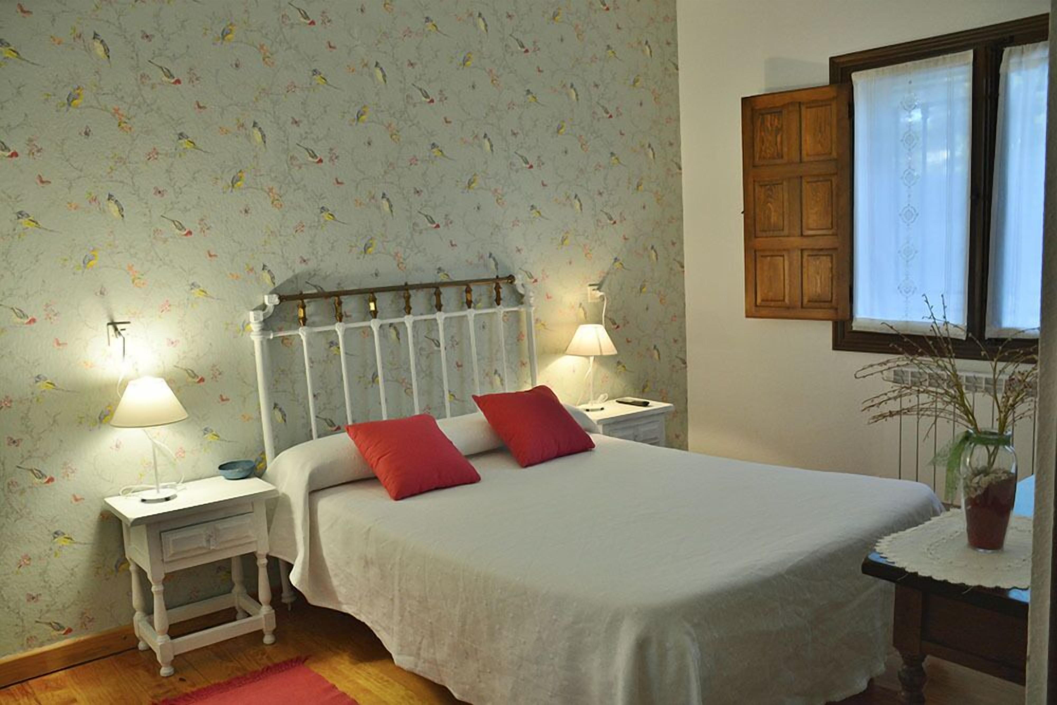 A bedroom with a bed, two nightstands, a window with curtains, and a wall with a floral pattern.