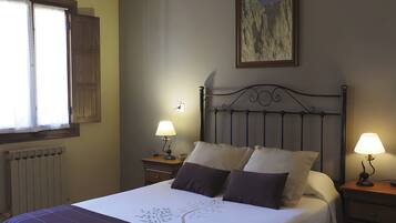 A neatly made bed with a white and purple bedspread, two bedside lamps, a wooden headboard, a painting on the wall, and a window with curtains.