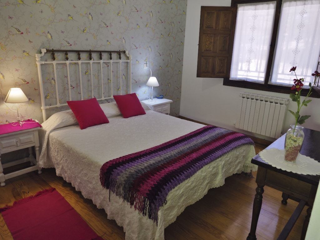 A bedroom with a bed, two bedside tables with lamps, a radiator, a window with curtains, and a red rug.