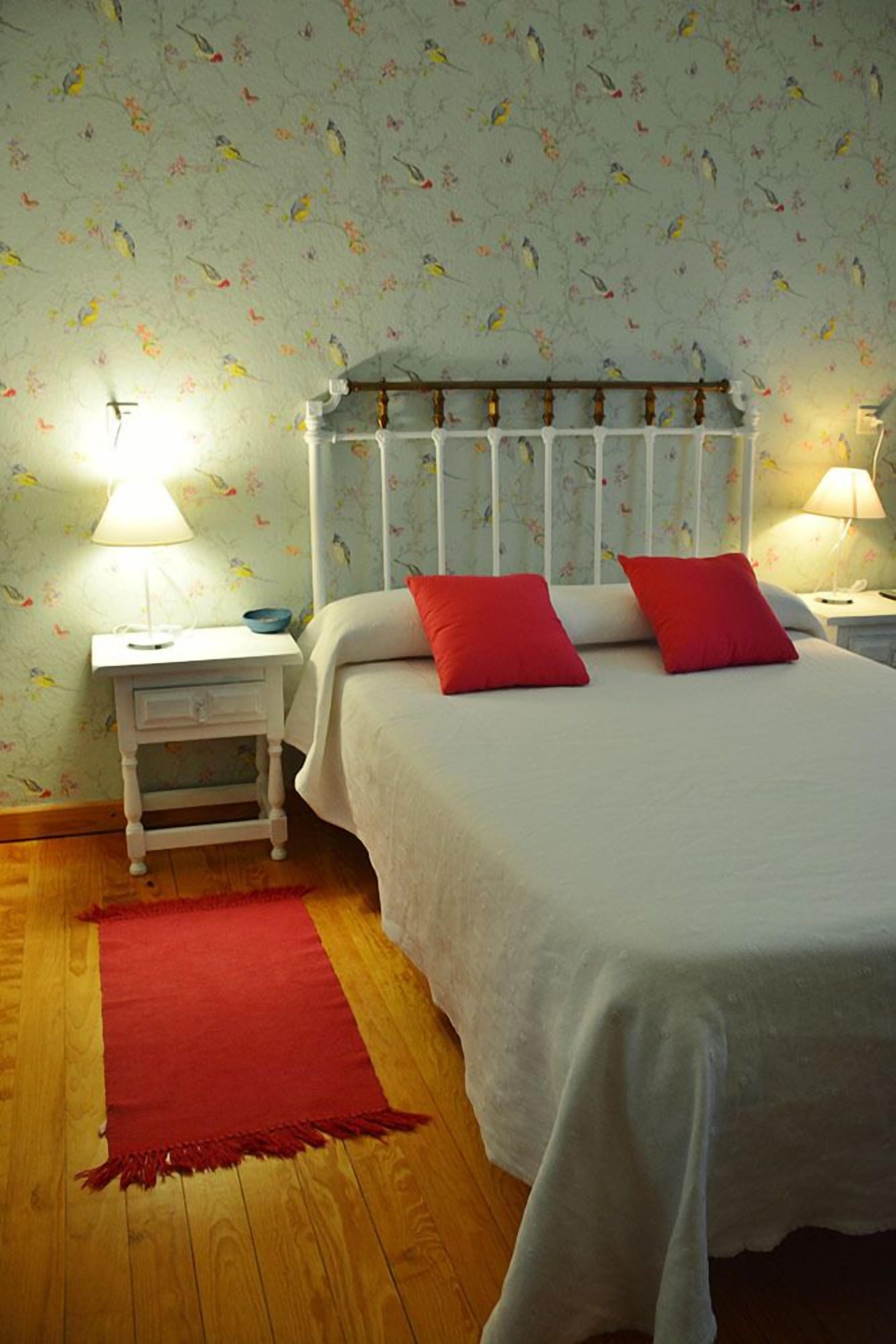 A bedroom with a bed, two red pillows, a white headboard, a nightstand with a lamp, and a red rug.