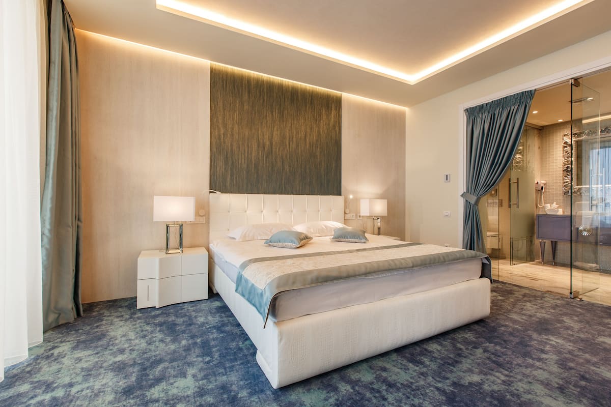 A modern hotel room with a large bed, bedside tables, a glass shower enclosure, and a view of the city.