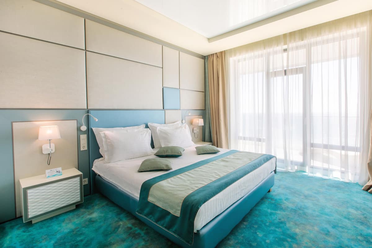 A modern hotel room with a large bed, a nightstand with a lamp, and a view of the outdoors through sheer curtains.