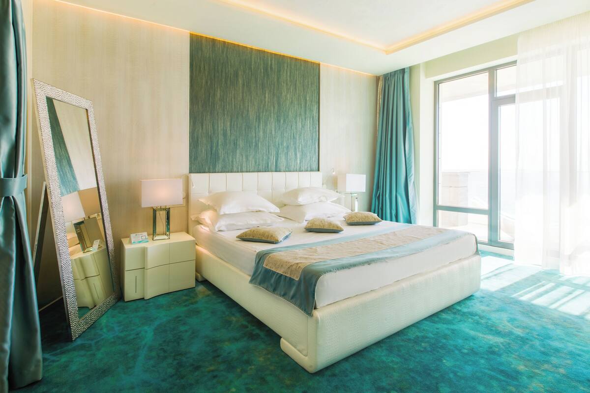 A hotel room with a large bed, a nightstand with a lamp, a mirror, and a view of the outdoors.
