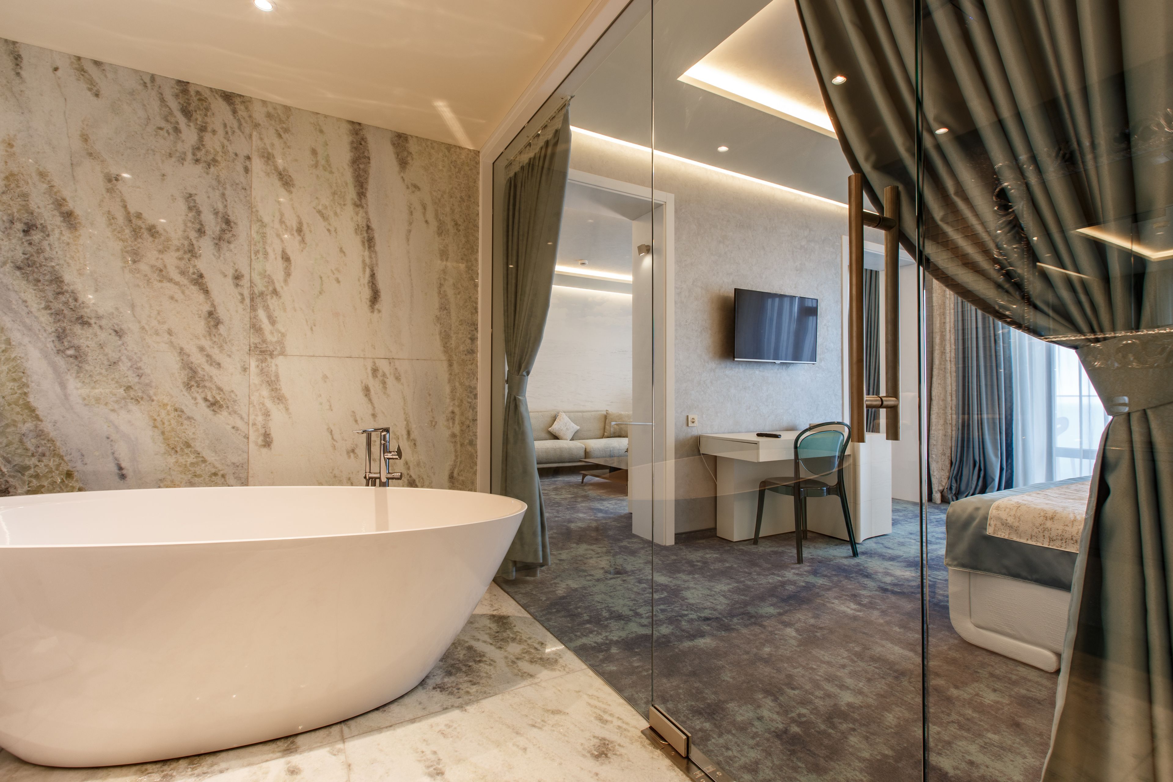 A modern bathroom with a freestanding bathtub, a glass shower enclosure, and a visible bedroom with a desk and chair.