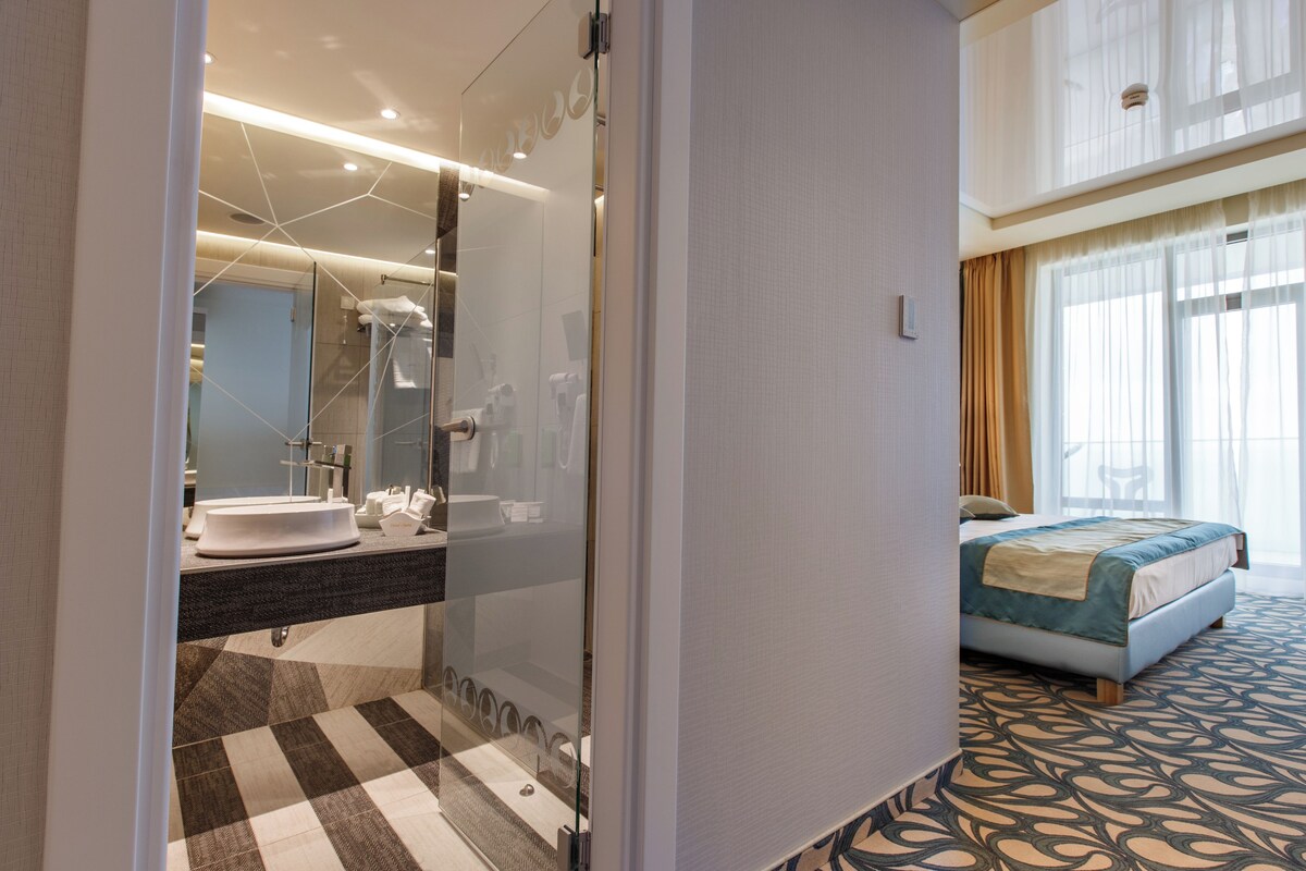 A modern hotel room with a large bed, a bathroom with a glass shower enclosure, and a checkered floor pattern.
