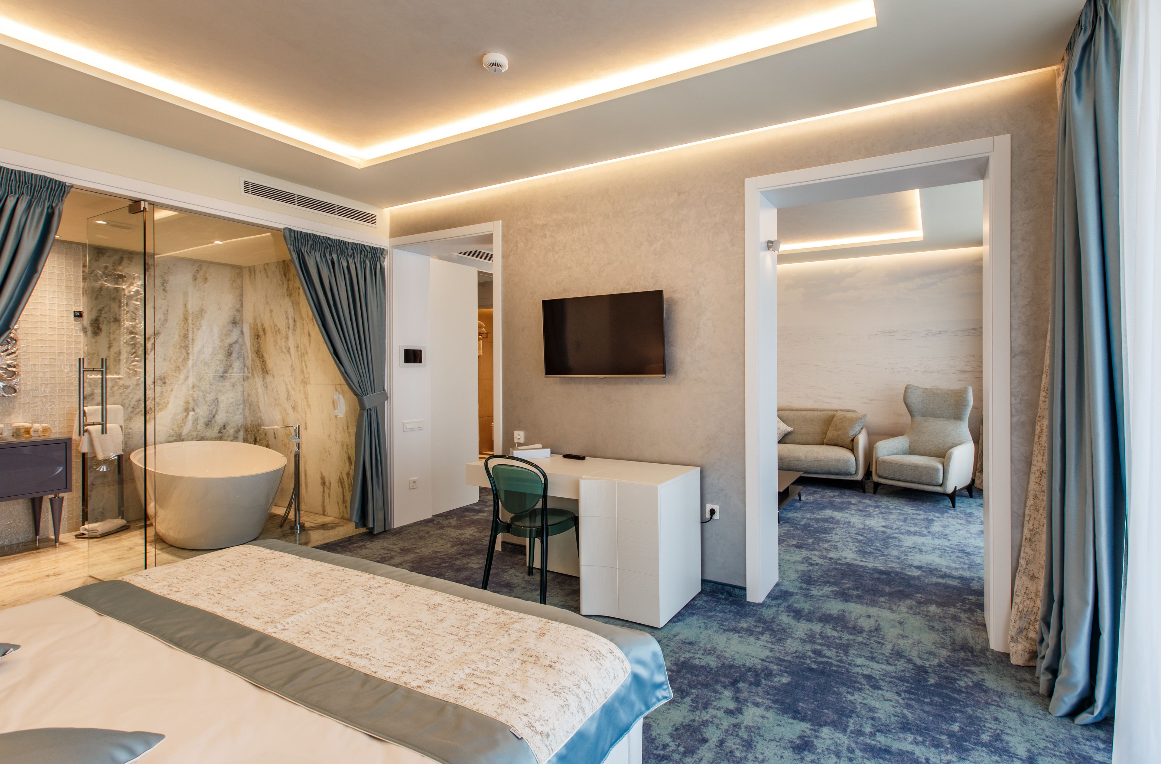 Habitación de hotel moderna con una cama grande, una zona de estar con escritorio y sillas, una bañera y un televisor montado en la pared.