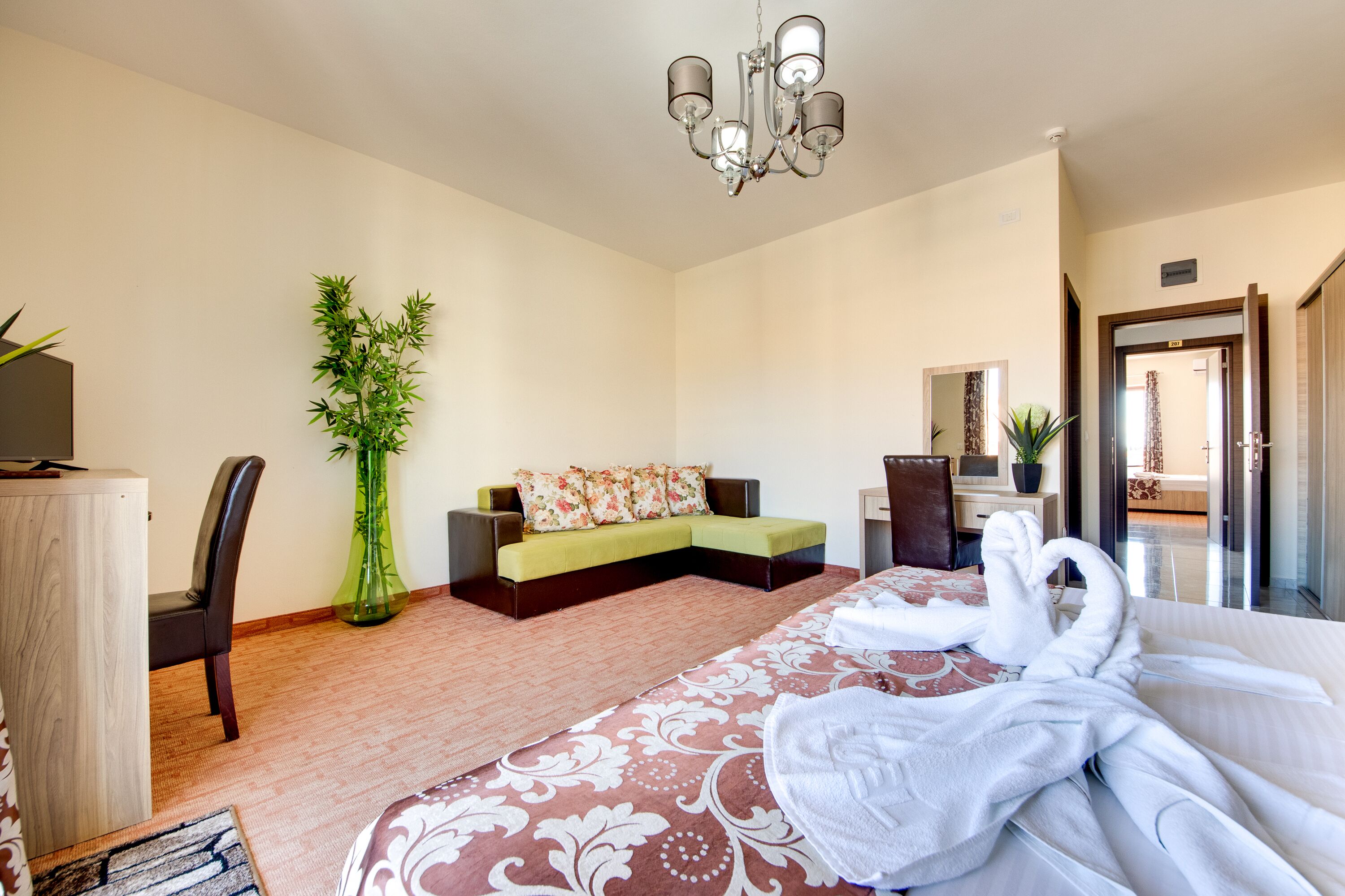 A hotel room with a bed, a sofa, a desk, a chair, a green plant, and a chandelier.