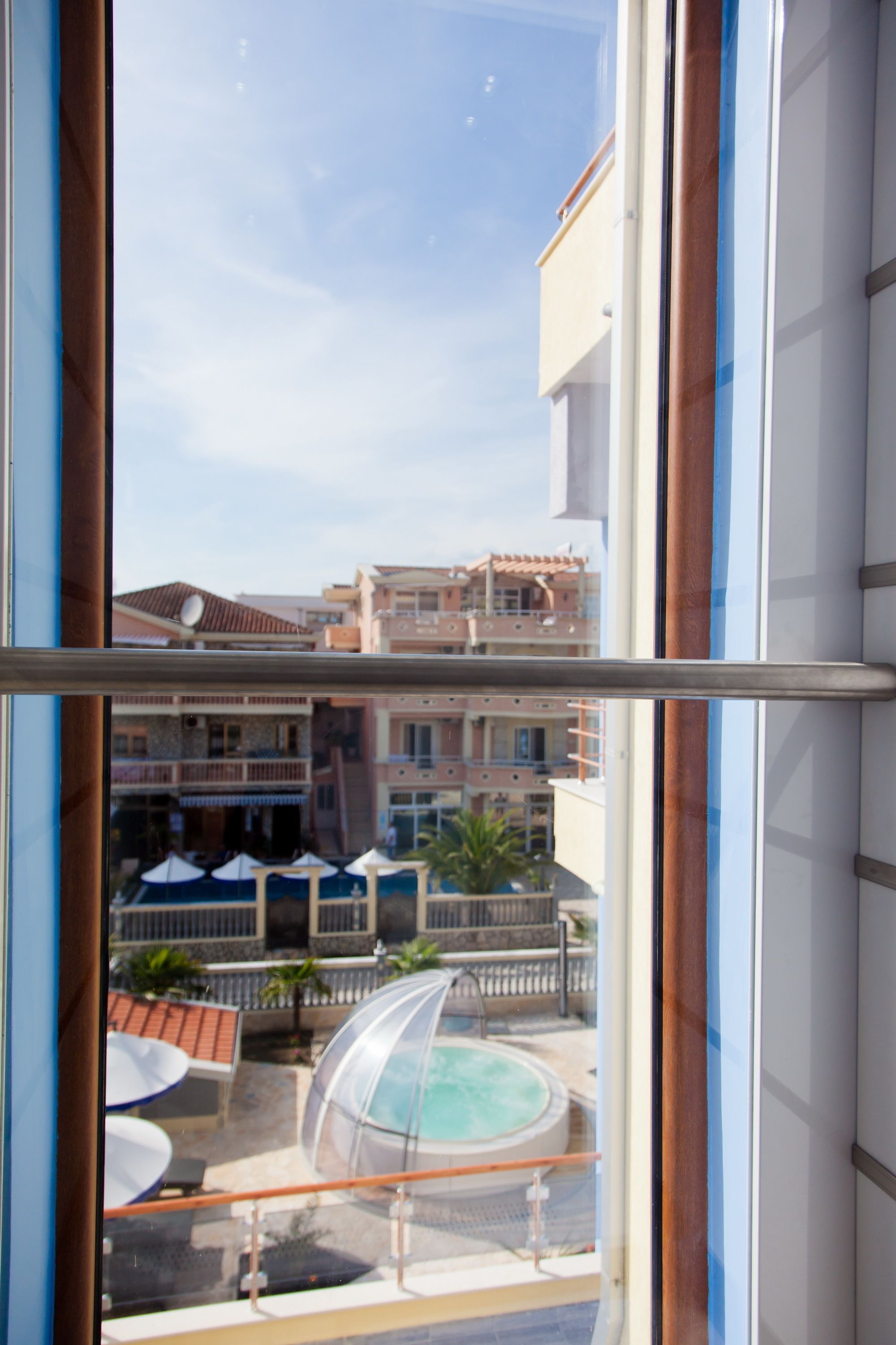 Vista a través de una ventana de un hotel con zona de piscina, sombrillas exteriores y un edificio con balcones.