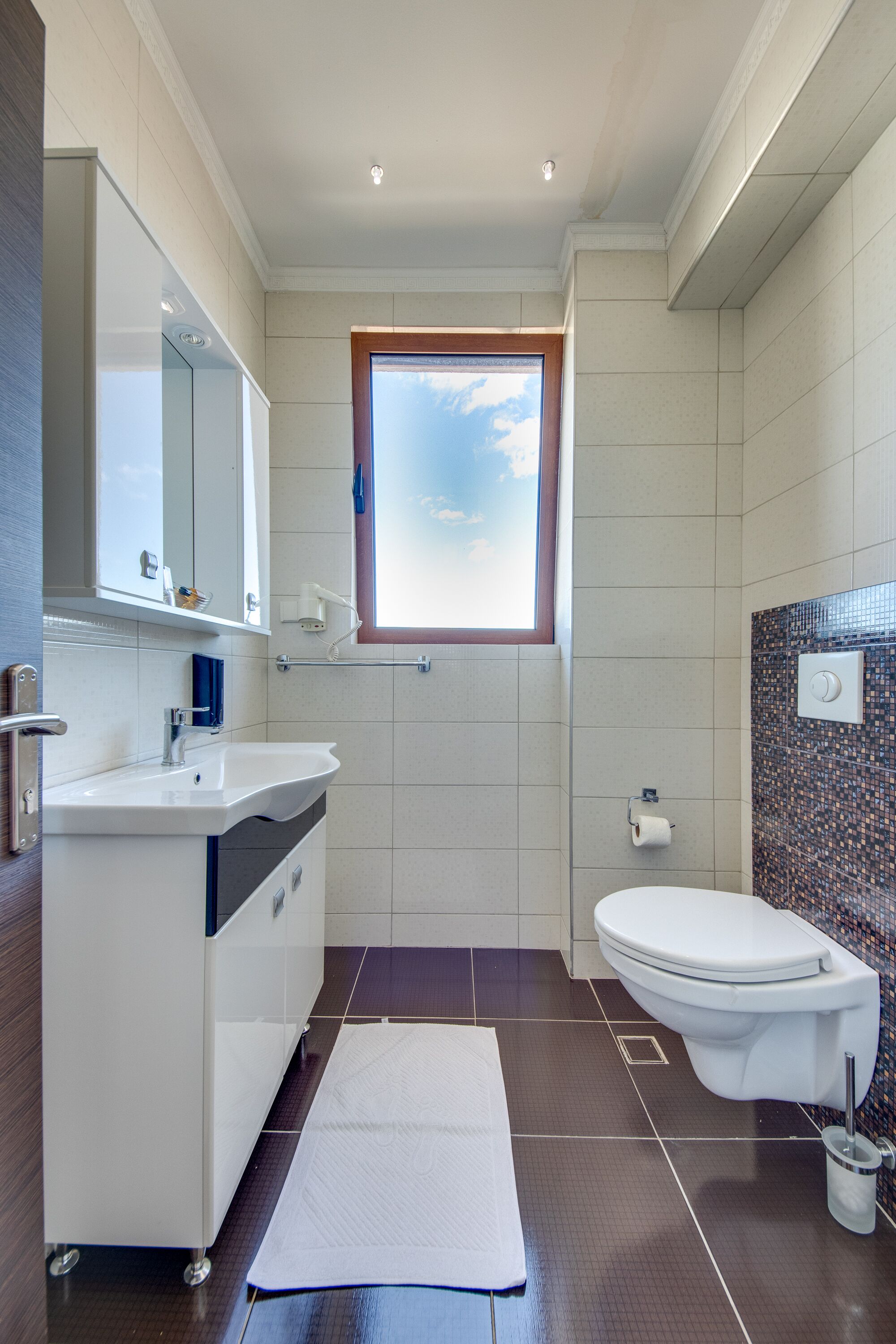 Un baño moderno con un lavamanos, espejo y retrete blancos. Cuenta con una ventana que proporciona luz natural y un estante pequeño sobre el lavamanos.