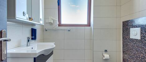 Un baño moderno con un lavamanos, espejo y retrete blancos. Cuenta con una ventana que proporciona luz natural y un estante pequeño sobre el lavamanos.