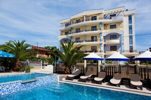 Un hotel con zona de piscina, sillones de descanso, sombrillas y un edificio de varios pisos con balcones.