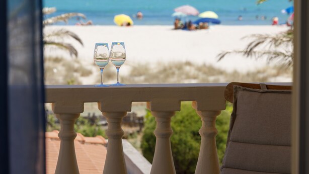 Un balcón con dos vasos de agua, con vistas a una playa con sombrillas y gente.