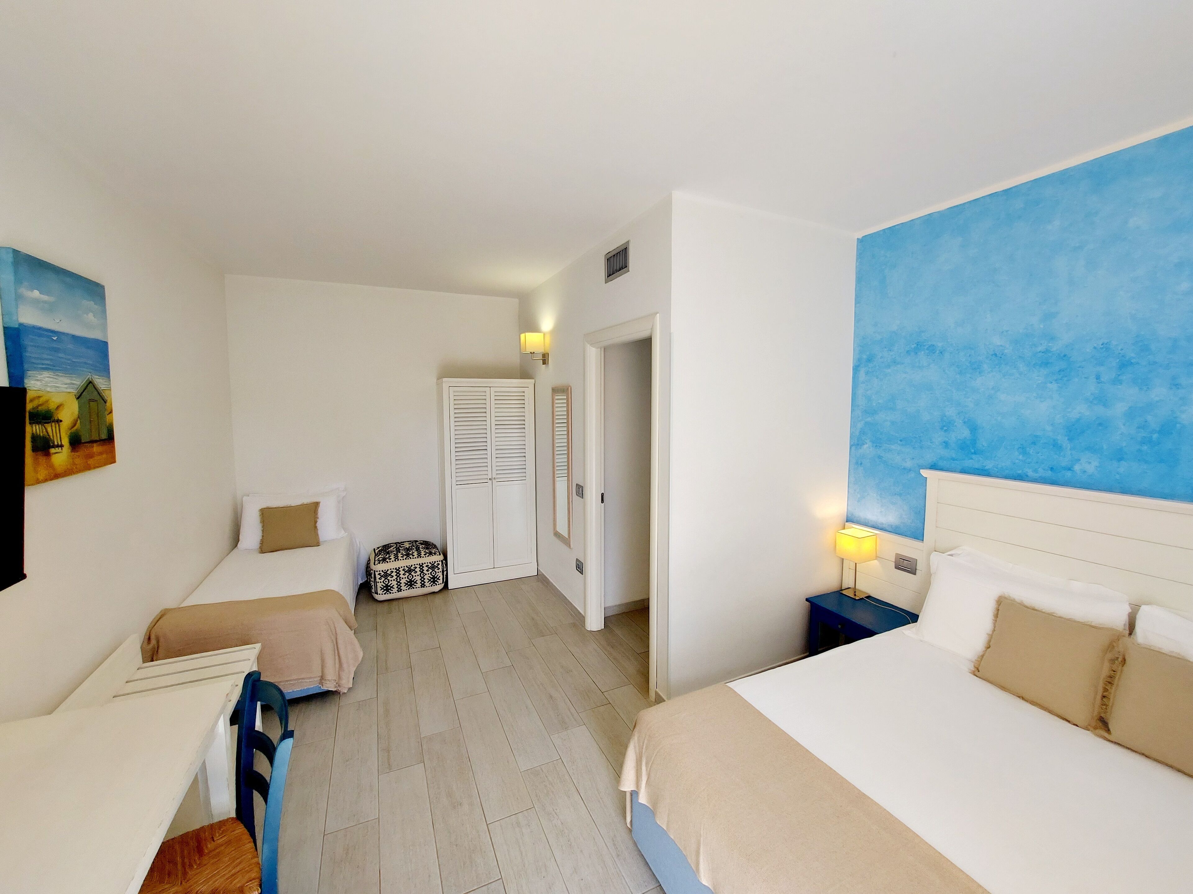 A hotel room with two beds, a small table, a chair, a wardrobe, and a painting on the wall.