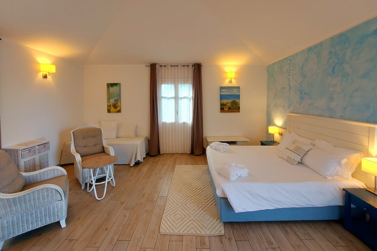 Hotellihuone, jossa on sänky, kaksi nojatuolia, pieni pöytä ja ikkuna, jossa on verhot.