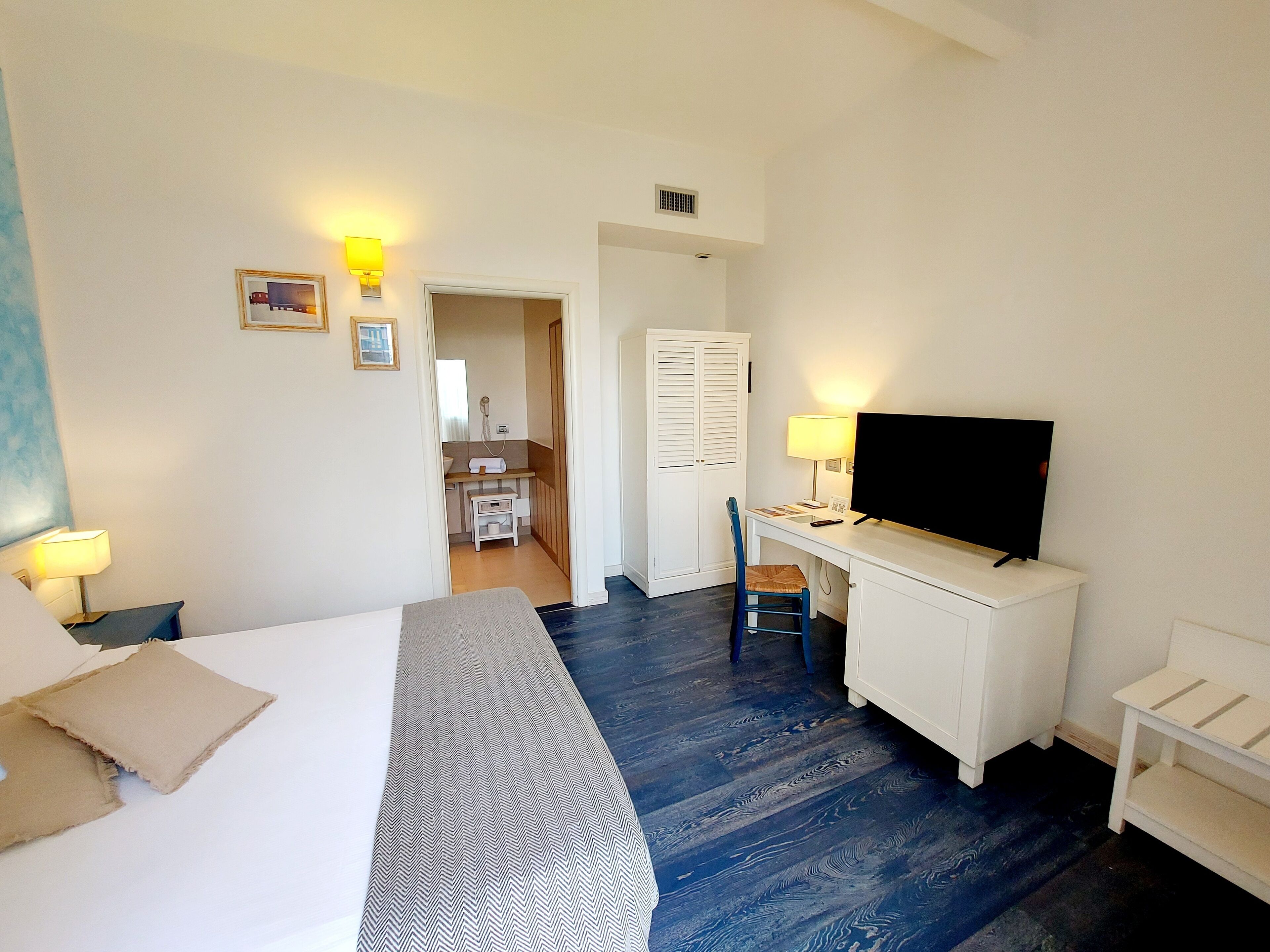 Une chambre d’hôtel avec un lit, un bureau avec une télévision, une chaise et une petite table. On peut apercevoir la salle de bain à travers une porte ouverte.