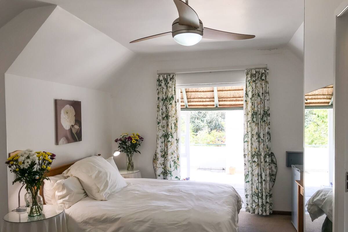 A bedroom with a bed, a bedside table with a lamp, a window with curtains, and a ceiling fan.