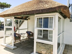 A covered patio area with a thatched roof, wicker furniture, and a view of a house in the background. - Howard's End Manor (Cape Town)