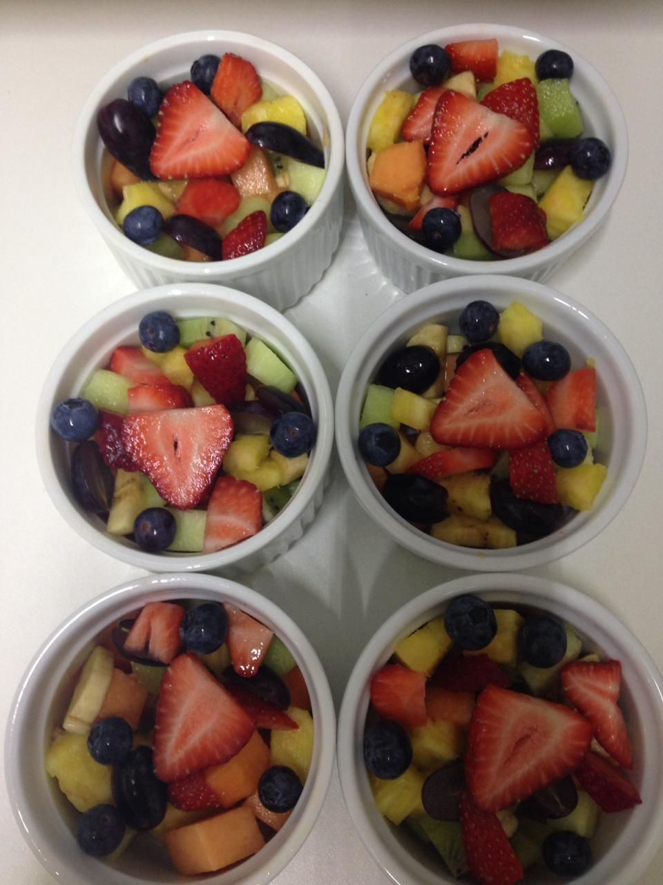 Six tasses blanches disposées en deux rangées de trois, chacune remplie d’une variété de fruits frais, notamment des fraises, des myrtilles et du melon.