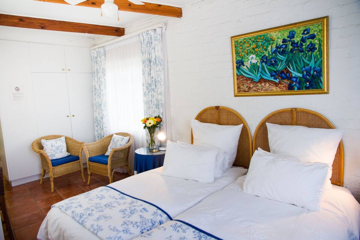 A bedroom with a double bed, two wicker chairs, a small table with a vase of flowers, and a painting on the wall.