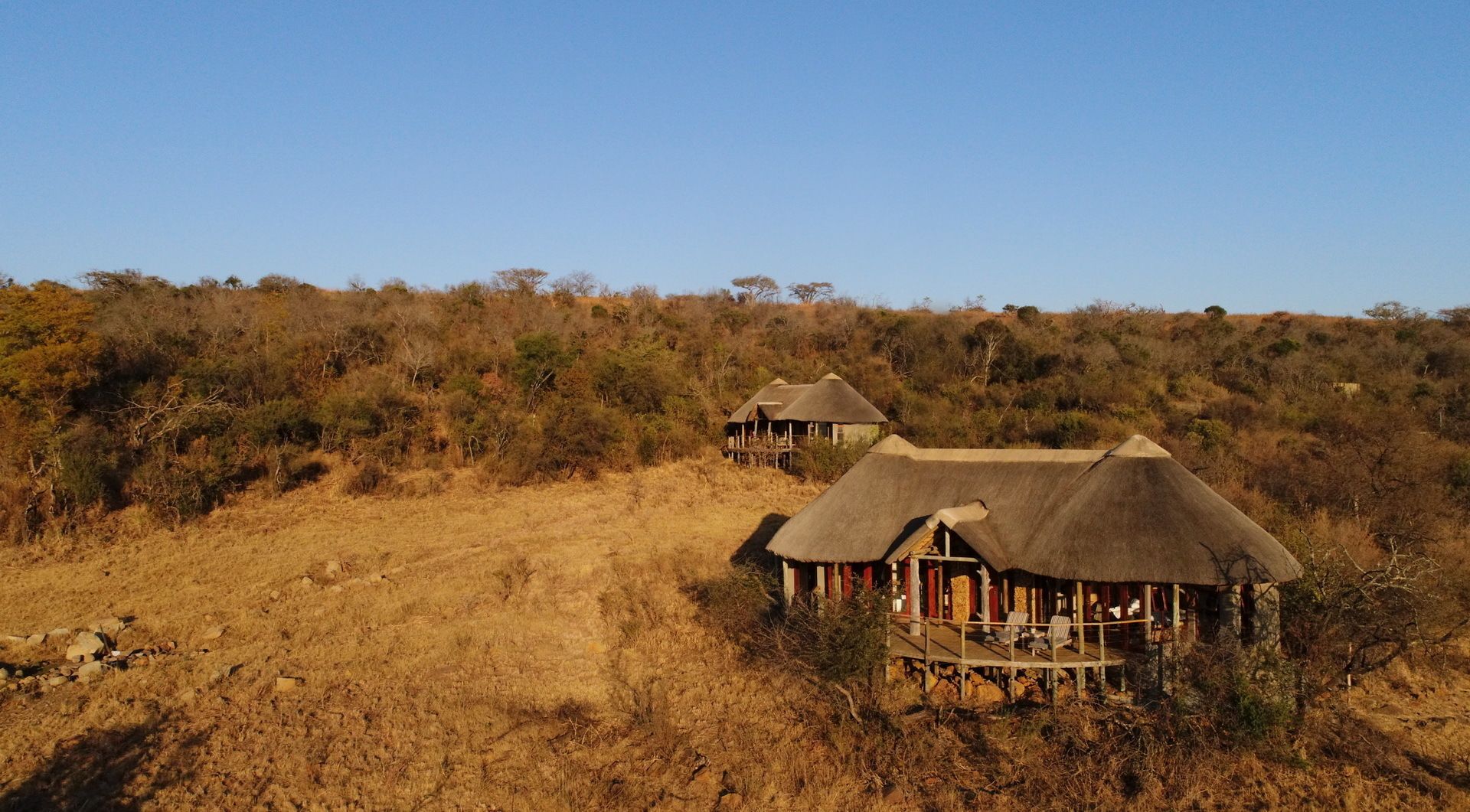 A thatched-roof structure with a wooden deck and seating area, surrounded by dry grassland and sparse trees.