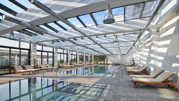 Una amplia piscina interior con techo iluminado por el sol y sillones de descanso a los lados.
