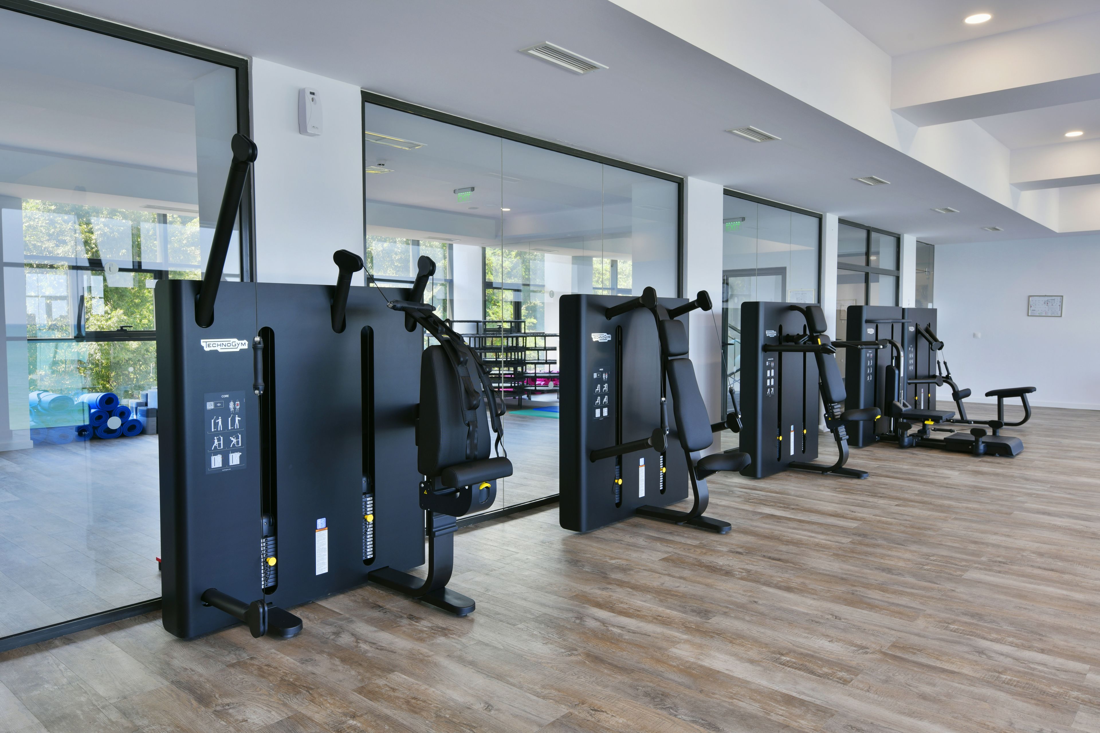 Un gimnasio moderno con máquinas de ejercicio, ventanas amplias y suelo de madera.