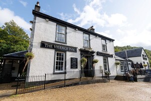 Um pub tradicional com uma placa que diz 'The Village Inn' e uma placa menor anunciando 'Pub & Restaurante Tradicional com Quartos'.