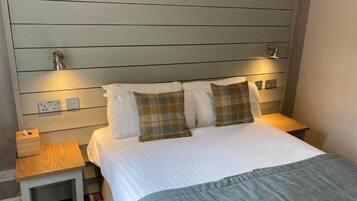 A hotel room with a bed, bedside tables, a nightstand, a lamp, and a power outlet.