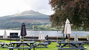 Picknicktische im Freien mit Sonnenschirmen, ein Schild mit der Aufschrift 'The Village Inn' und ein See mit einem Berg im Hintergrund.