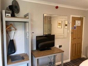 Une chambre d’hôtel avec une télévision posée sur une table en bois, un miroir au mur, une porte avec une poignée rouge et un ventilateur suspendu au plafond.